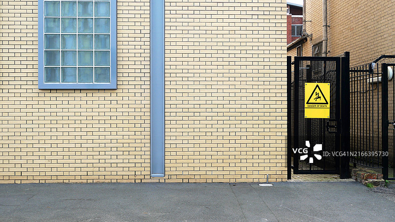 黄色砖砌工业建筑的外立面特写,带有盲窗、电击危险标志的门和铁栅栏。沥青街道。伦敦,英格兰,联合王国。阳光。自然色彩。图片素材