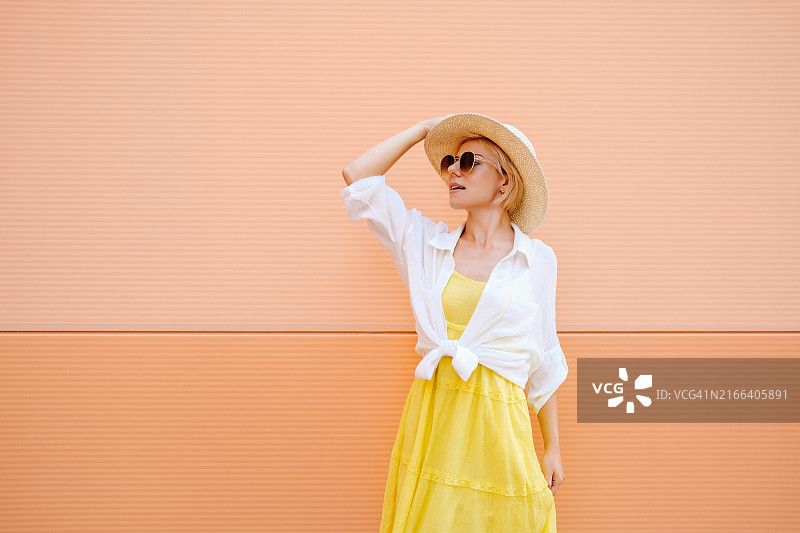 年轻女子身穿衬衫、连衣裙，头戴宽边帽，站在桃色墙壁旁图片素材