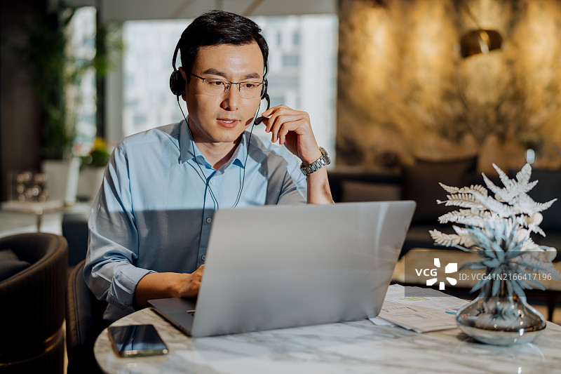 在现代办公室里戴着耳机参加视频会议的亚洲商人，捕捉严肃的表情。图片素材