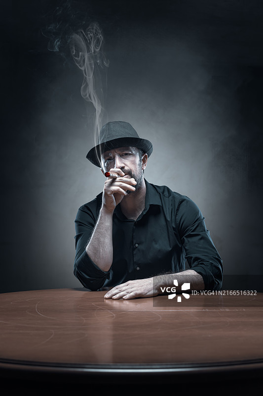 在暗室里戴着帽子抽着雪茄坐在桌子旁的黑帮老大图片素材
