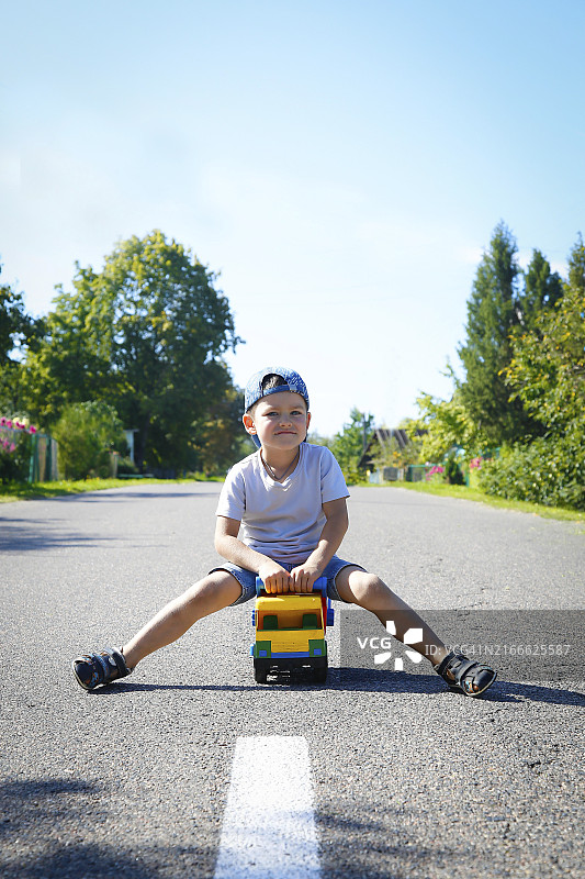 阳光明媚的日子里，男孩坐在马路中央的玩具卡车上，周围环绕着树木图片素材