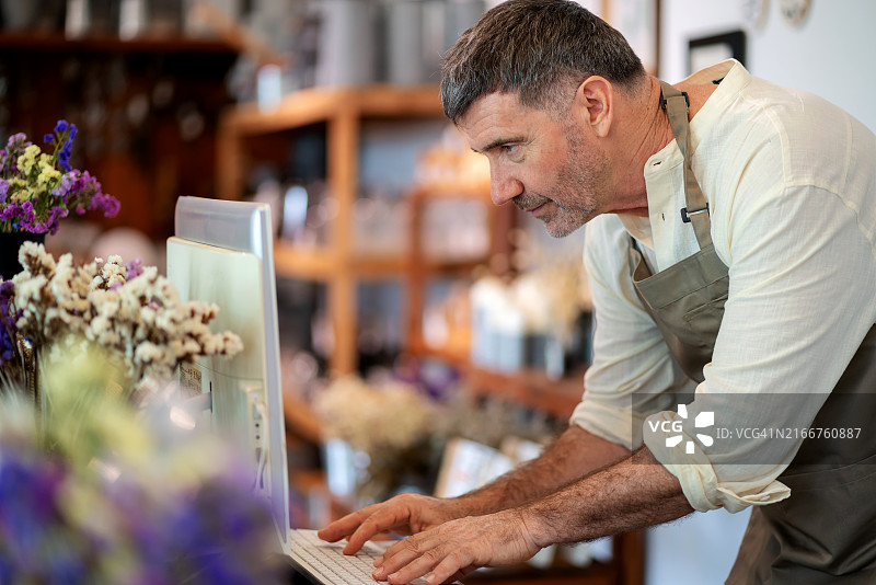 经验丰富的老员工站在家居装饰店的收银台前，用电脑管理库存和交易，身穿围裙，热情地与顾客互动，提供建议和解决方案图片素材