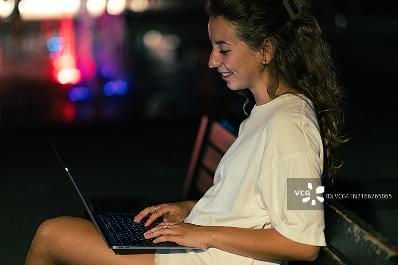 年轻女子深夜在城市长凳上使用笔记本电脑图片素材