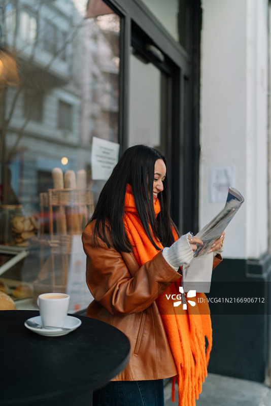 穿着橙色外套的年轻女子在户外咖啡馆看报纸喝咖啡图片素材