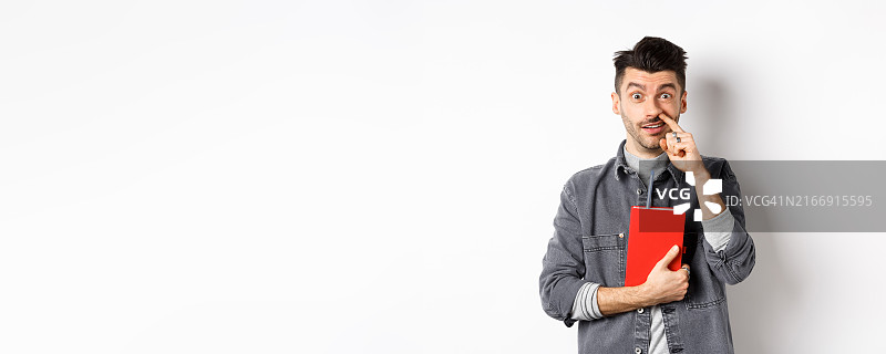 对着镜头抠鼻子且拿着书的年轻男人，看起来很傻，站在白色背景前图片素材