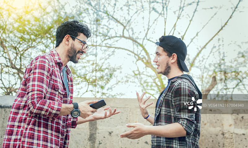 两个朋友微笑着用手机聊天。两个青少年面对面交谈，两个年轻的朋友在户外聊天图片素材