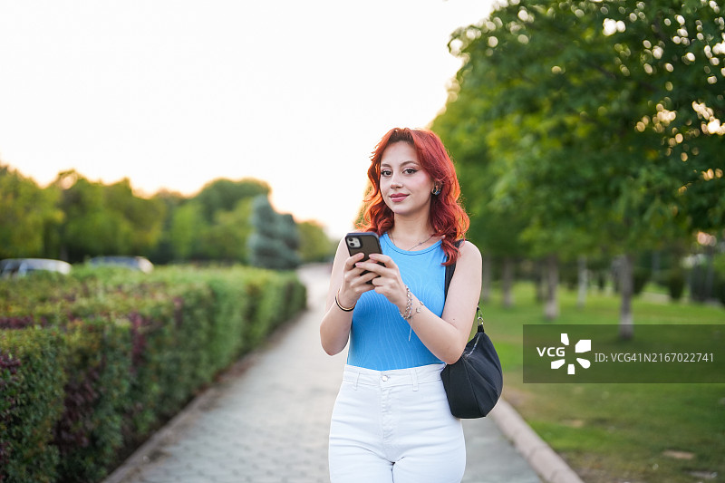 在开阔的田野里看手机的年轻红发女子图片素材