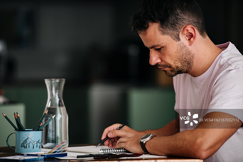 一位穿着淡紫色T恤、戴着皮表带手表的年长白人男子在木桌上用绿色铅笔画素描图片素材