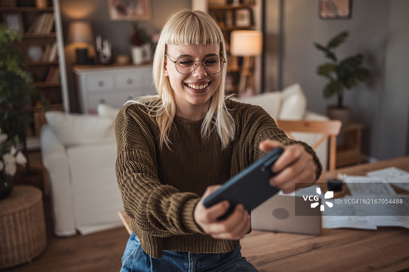 快乐的白人女孩在客厅的桌旁用智能手机玩在线视频游戏，放松身心图片素材