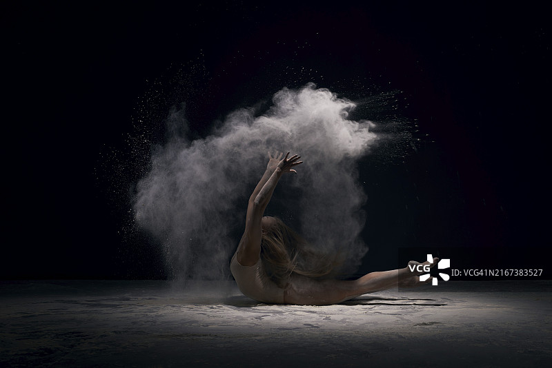 女美丽体操运动员，她的金发宽松的米色紧身衣优雅地弯曲在黑色的背景与白色灰尘全身拍摄的侧面图片素材