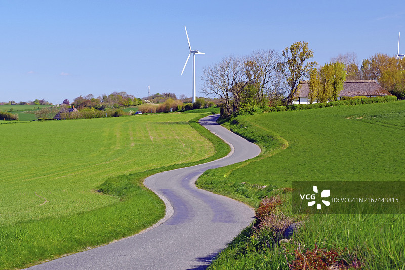 一条蜿蜒的道路穿过绿色的田野，背景是风力涡轮机，自行车旅游，庄园路线，丹麦朗厄兰岛诺雷博勒，欧洲图片素材