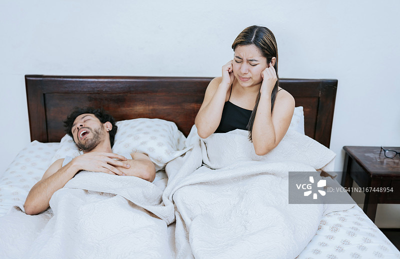 丈夫打鼾，妻子捂着耳朵难受。睡眠呼吸暂停概念，卧室里打鼾的男人和捂着耳朵的妻子图片素材