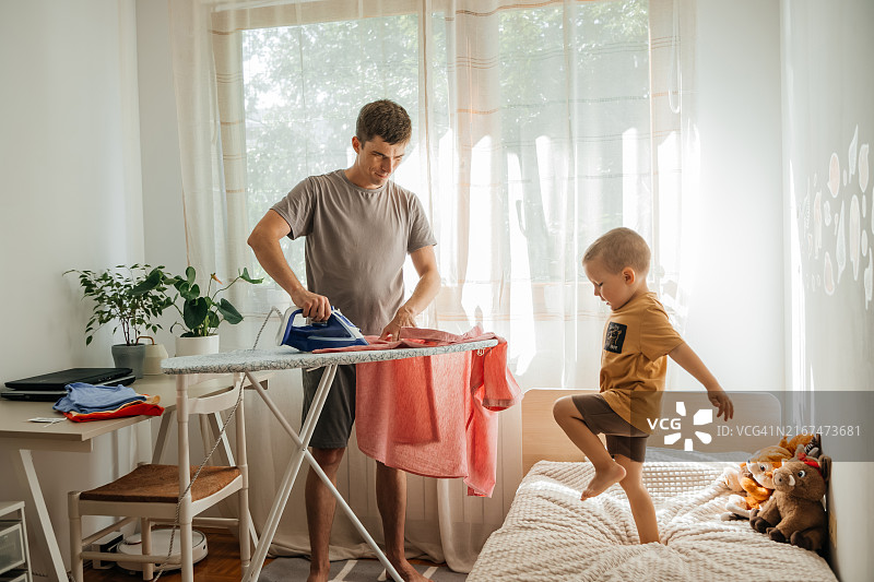 居家生活：男人休产假照顾孩子并做家务，熨烫衣服，孩子分散了父亲的注意力图片素材