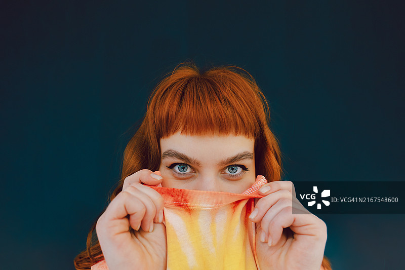 俏皮地躲藏在他的T恤后面的富有表现力的红发年轻女人图片素材