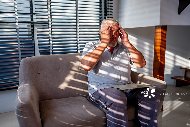 老年男子坐在明亮的客厅里，舒适的椅子上使用笔记本电脑，窗帘拉开。图片素材