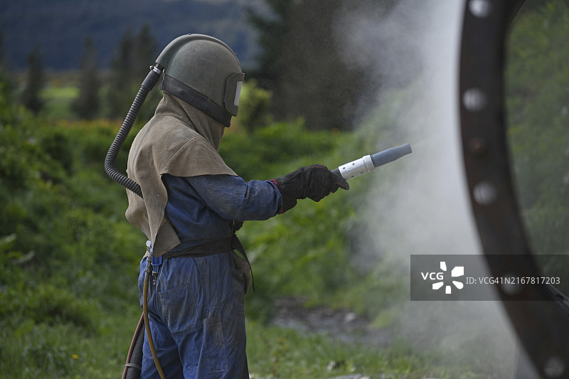 穿着全套安全装备的男人用玻璃砂喷砂处理肉类干燥机的钢制外壳图片素材