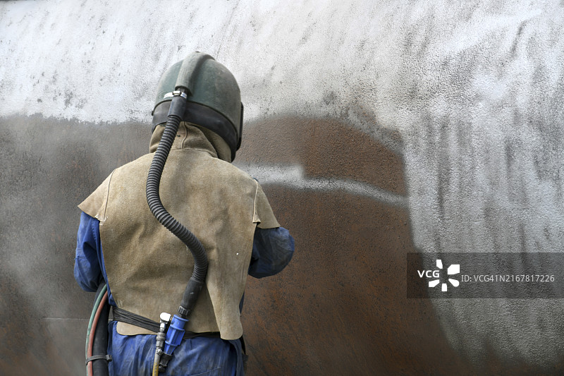 一名男子穿着全套安全装备，用碎玻璃喷砂处理渲染厂肉类干燥机的钢制外壳图片素材