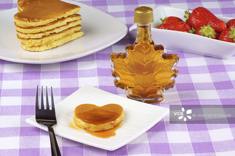 迷你心形煎饼，白色盘子上淋有糖浆，配有叉子。背景是一叠煎饼、一瓶枫糖浆和一些草莓，是情人节的完美早餐图片素材