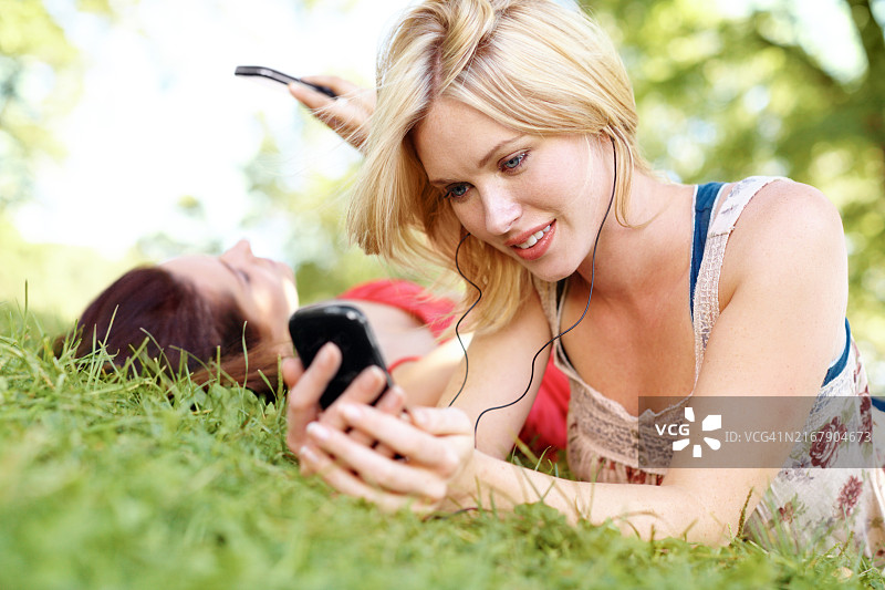音乐，手机，与女性朋友在草地上放松，在夏季周末一起播放。耳机，互联网和公园里的人们躺在绿色的草坪或地上，享受联谊和休闲图片素材