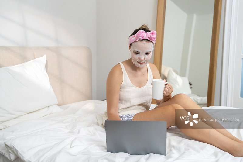在家里戴着美容面膜躺在床上享受使用笔记本电脑的女人图片素材