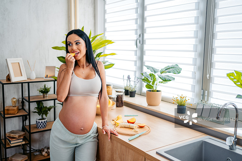 怀孕的中年妇女在家里的厨房吃切好的水果图片素材