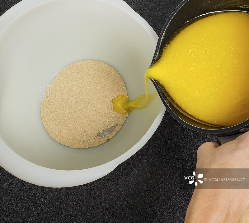 手将融化的黄油从黑色水壶倒入黑色桌子上装有干酵母的白色碗中图片素材