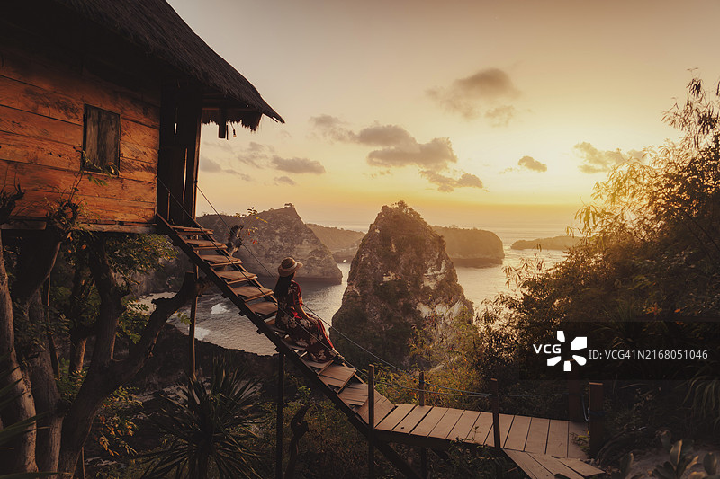 戴着草帽、穿着红色碎花裙的女人站在树屋的木制台阶上，凝视着印度尼西亚巴厘岛努沙 penida 令人惊叹的日出图片素材