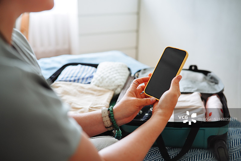 一位加大码女性手中拿着黑屏智能手机，身后放着打包好的行李箱，代表旅行计划图片素材