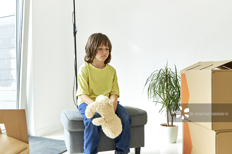 悲伤的男孩抱着毛绒玩具坐在新房的脚凳上图片素材