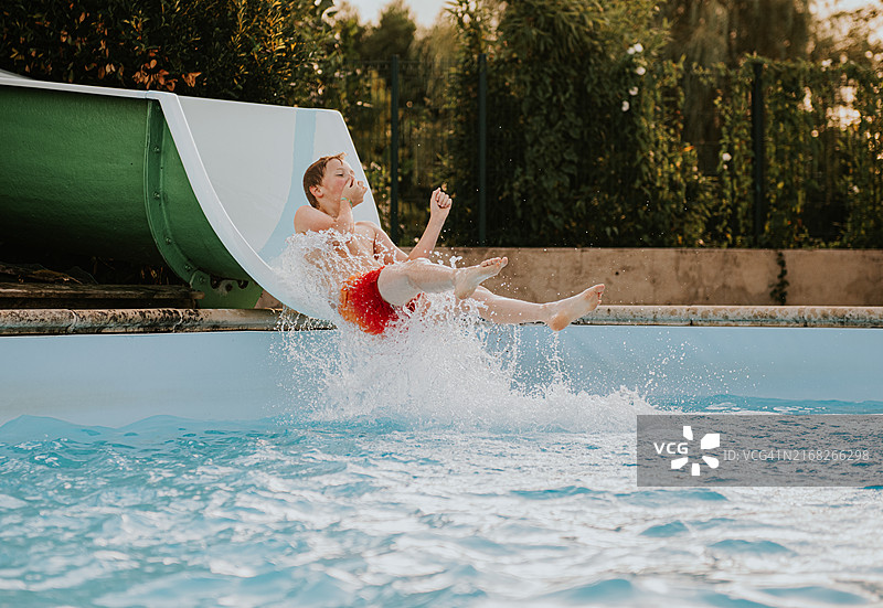 一个快乐的孩子在阳光明媚的下午，从水滑梯滑入波光粼粼的游泳池——当他准备迎接冲击时，他捏住了鼻子图片素材