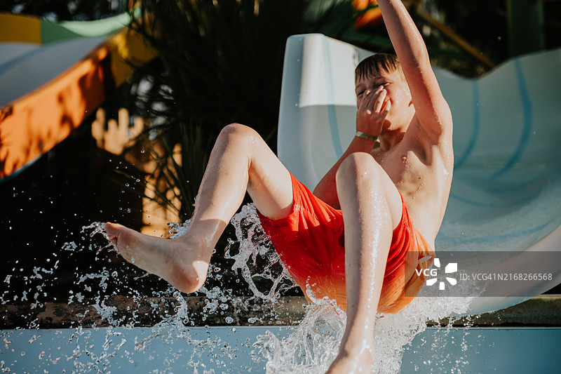 一个快乐的孩子在阳光明媚的下午，从水滑梯滑入波光粼粼的游泳池——当他准备迎接冲击时，他捏住了鼻子图片素材