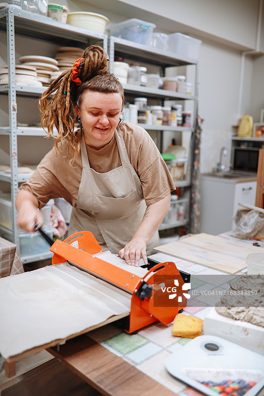 体型偏胖的女陶艺家在工作室用滚动机为盘子制作光滑的陶土层图片素材