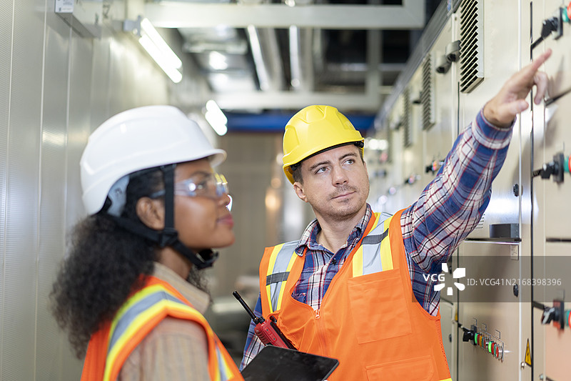 在电气控制室里，穿着橙色安全服的准女工程师和男同事图片素材