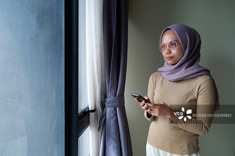 戴着头巾的年轻穆斯林女人在客厅窗边使用智能手机图片素材