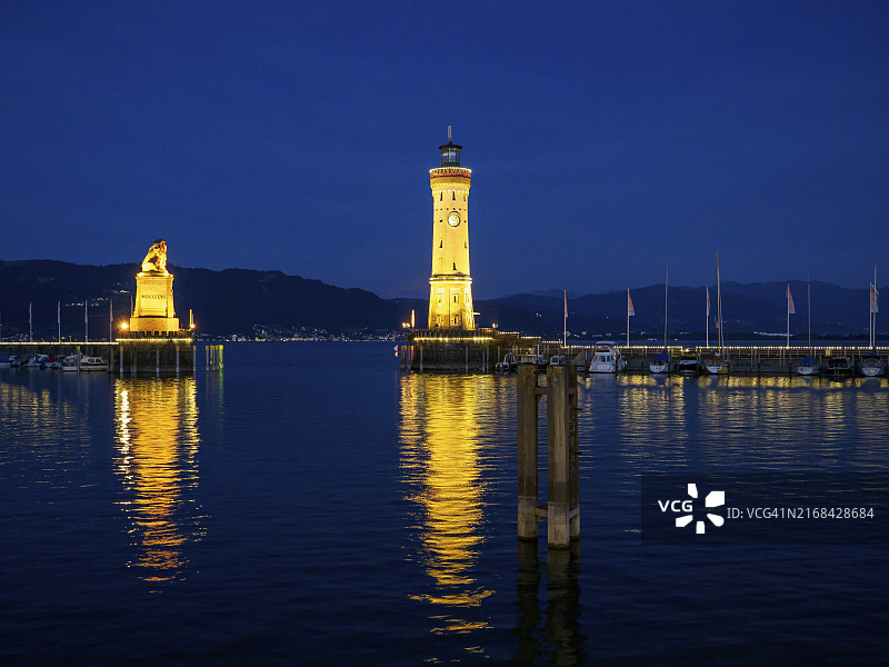 夜晚，灯塔和雕像在平静的港口水中倒映着山景，德国巴伐利亚林道图片素材