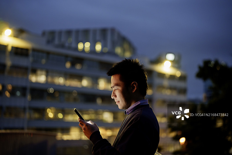 专注的亚洲男性在灯火通明的城市背景下阅读智能手机图片素材