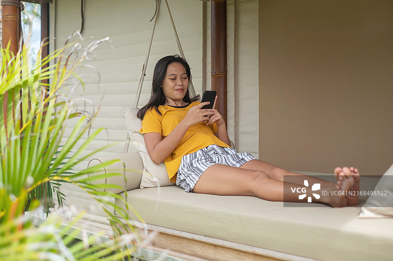 在门廊秋千上玩手机游戏的印尼年轻女人图片素材