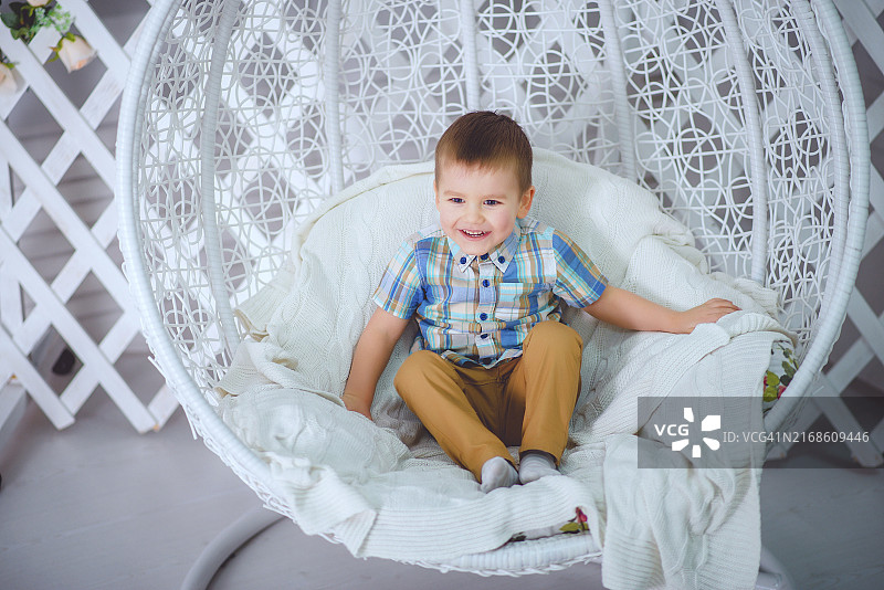 穿着格子衬衫和黄色裤子的男孩坐在白色编织椅子上微笑图片素材