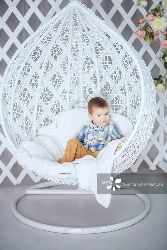 一个婴儿舒适地坐在白色柳条椅上，穿着格子衬衫和米色裤子，背景是装饰着鲜花的格子图片素材