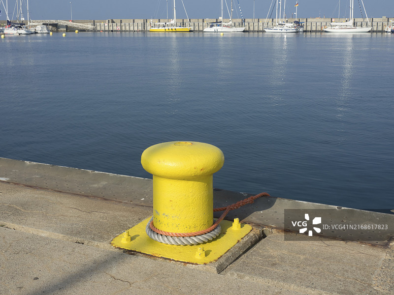 德国黑尔戈兰岛港口码头边缘的黄色系缆桩，平静的水面，背景是帆船图片素材