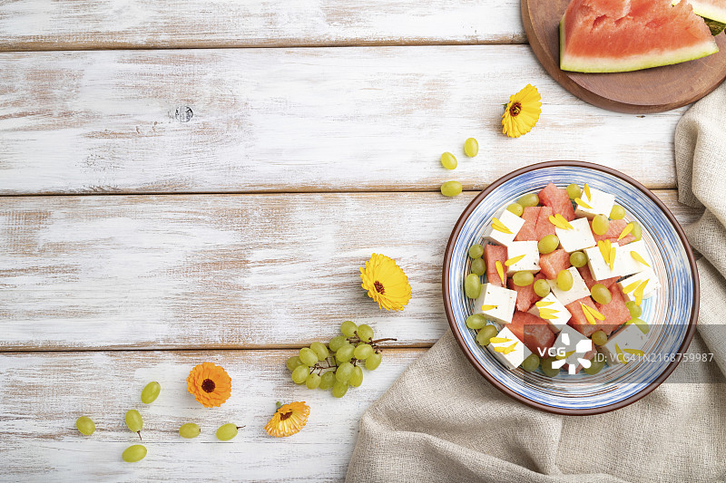 素食沙拉：西瓜、羊乳酪和葡萄，蓝色陶瓷盘，白色木质背景和亚麻纺织品。图片素材