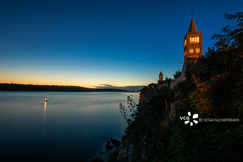 日落后漂浮在海上的灯光渔船，海岸边的教堂塔楼图片素材