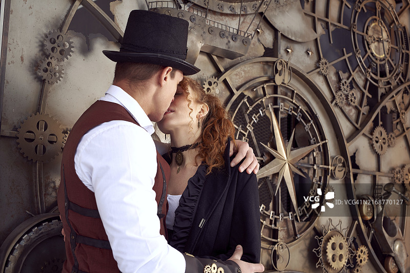 年轻的红发女人和男人穿着老式蒸汽朋克风格的服装站在旧机械旁边亲吻图片素材