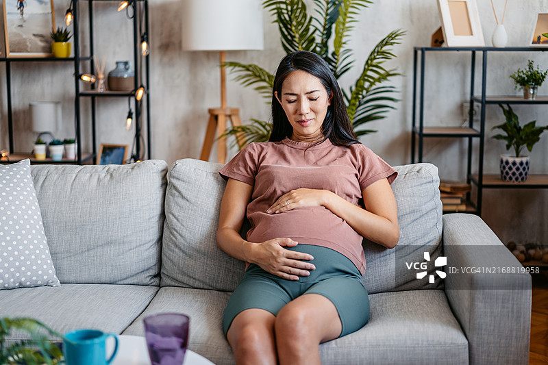 中年怀孕亚洲女性坐在客厅沙发上，感到胃痛图片素材