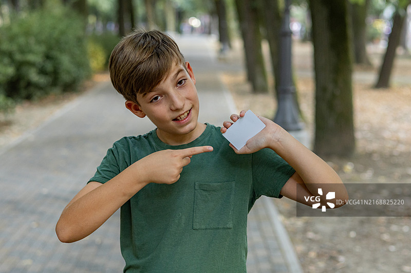 可爱的男孩在公园外摆姿势，手里拿着他的借记卡。快乐的男孩向镜头展示一张空白的银行卡。图片素材