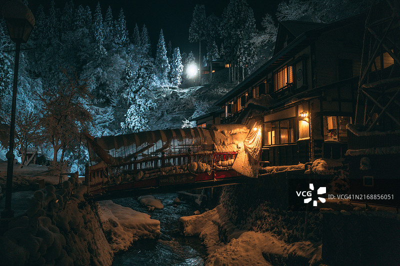 冬季，日本山形县尾花泽市的银山温泉被白雪覆盖，木制建筑和灯笼照亮的街道两侧是银山河图片素材