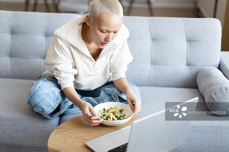 在家坐在沙发上吃沙拉并使用笔记本电脑工作的年轻亚洲女性图片素材
