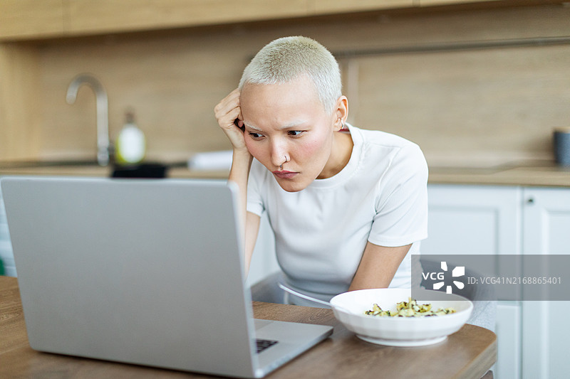 年轻女子坐在厨房里，一边吃东西一边用笔记本电脑工作图片素材