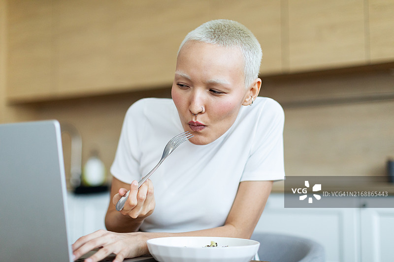 年轻女子坐在厨房里，一边吃东西一边用笔记本电脑工作图片素材