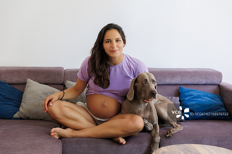 孕妇坐在客厅沙发上拥抱她的狗，对着镜头微笑图片素材
